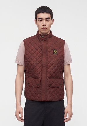Junger Mann trägt eine ärmellose braune gesteppte Weste mit Vordertaschen und einem Logopatch über einem hellrosa kurzärmeligen Hemd.