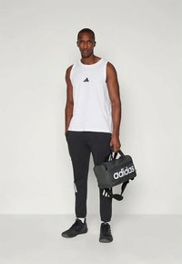 Mustat urheiluhousut, valkoinen hihaton toppi mustalla logolla ja mustat lenkkarit. Pitelee harmaata kuntosalilaukkua, jossa on valkoista brändäystä.