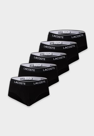 Homme torse nu portant un slip noir Lacoste, debout pieds nus sur un fond blanc uni, les mains dans le dos.