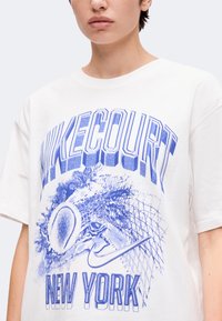 Personne portant un T-shirt blanc avec du texte graphique bleu "NIKE COURT" et "NEW YORK" et une illustration d’une balle de tennis frappant un filet.