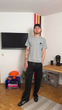 Harma t-paita, jossa on pieni sininen logo, mustat verkkarit, mustat lenkkarit valkoisilla raitoilla ja musta lippis. Seisoo hyvin sisustetussa huoneessa.