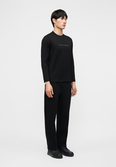 Άνδρας που στέκεται με μαύρο μακρυμάνικο πουκάμισο με διακριτικό λογότυπο, μαύρο παντελόνι και μαύρα αθλητικά παπούτσια, μπροστά σε απλό φωτεινό φόντο.