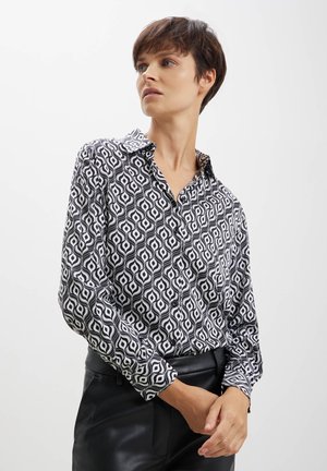 Blusa bianca e nera con un motivo geometrico, colletto, maniche lunghe e texture setosa, abbinata a pantaloni neri.