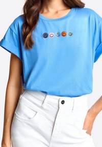 Blå kortärmad t-shirt med fem färgglada broderade leende ansikten på framsidan. Matchad med vita högmidjade byxor.