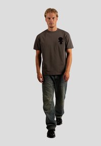 Camiseta de algodón gris con mangas cortas, que presenta una figura de dibujos animados negra con un detalle de corazón rojo en el pecho, combinada con vaqueros azul claros.