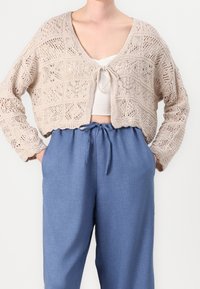 Beige kort stickad kofta med öppet mönster och frontknytning. Kombinerad med ljusblå avslappnade byxor med dragsko i midjan.