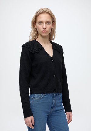 Jeune femme aux cheveux blonds ondulés portant un cardigan noir boutonné avec un grand col en dentelle et un jean bleu, debout devant un fond blanc.