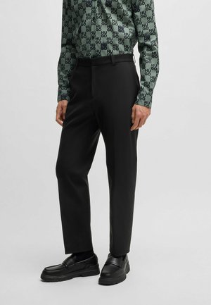 Pantaloni neri su misura con taglio dritto, indossati con una camicia verde a maniche lunghe con fantasia e mocassini neri robusti.