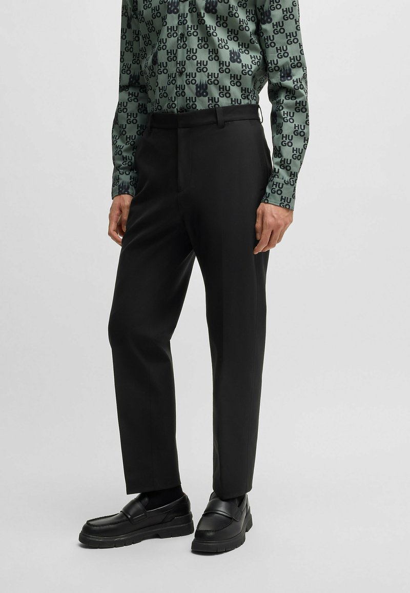 Pantaloni neri su misura con taglio dritto, indossati con una camicia verde a maniche lunghe con fantasia e mocassini neri robusti.