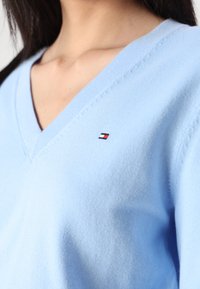 Lys blå strikket genser med V-hals og liten logo-patch i rød, hvit og blå nær halsområdet. Myk tekstur.
