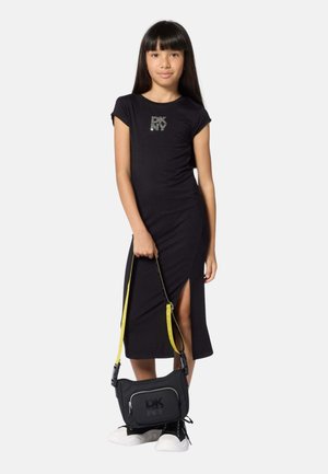 Meisje draagt een zwarte DKNY-jurk met een zijsplit, houdt een zwarte DKNY-tas met gele band vast en draagt zwart-witte sneakers.