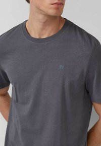 Mann in einem schlichten, dunkelgrauen, kurzärmeligen Rundhalsausschnitt-T-Shirt mit kleinem, dezentem Logo auf der linken Brust, stehend vor einem hellen Hintergrund.