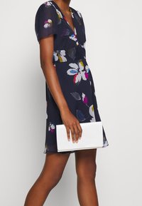 Blumenmusterkleid mit kurzen Ärmeln, tiefem V-Ausschnitt und dunkelblauem Hintergrund. Begleitet von einer rechteckigen weißen Clutch in der Hand.