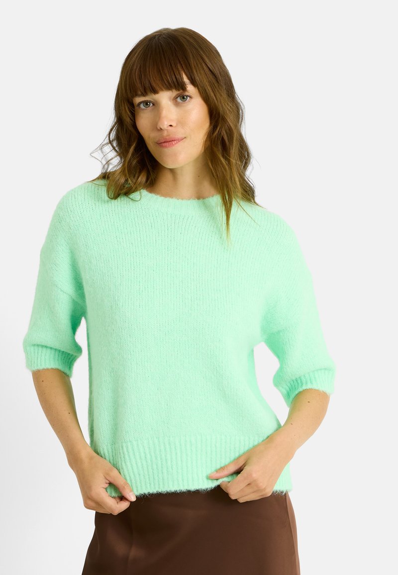 Pull en maille vert menthe à manches courtes, avec un col rond et un ourlet côtelé. Texture douce et coupe décontractée, associé à une jupe marron.