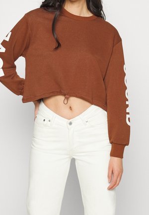 Woman wearing a cropped brown sweatshirt with white text on the sleeves and white high-waisted jeans, hand resting on hip.