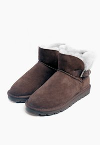Botines de ante marrón con un forro de piel de oveja blanca, de perfil bajo, con un detalle de hebilla lateral y una suela de goma texturizada para mayor agarre.