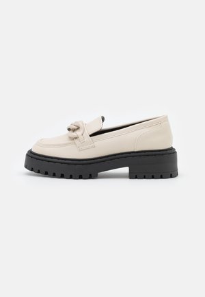 Mocassin beige avec une semelle épaisse noire à crampons et un détail de nœud décoratif sur le dessus, vu de côté sur fond blanc.