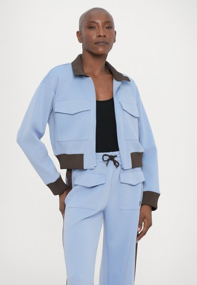 Jaqueta azul clara com fecho éclair, colarinho e punhos castanhos, apresentando dois bolsos frontais. Usada com calças azul claro combinando e uma blusa preta.