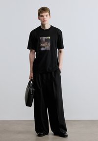 Μαύρο βαμβακερό μπλουζάκι με γραφικό σχέδιο που φέρει τη λέξη «HUGO». Συνδυασμένο με φαρδιά μαύρη παντελόνα και ματ μαύρη τσάντα.