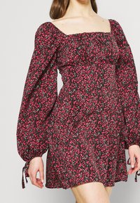 Musta mekko, jossa on vaaleanpunaisia kukkakuoseja, neliökaula-aukko, pitkät puffihihat ja nauhatyyliset yksityiskohdat hihoissa. Pehmeä kangas, jossa on korostettu vartaloa.