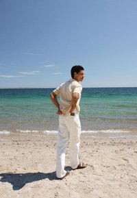 Άνδρας με κοντομάνικο πουκάμισο, λευκό παντελόνι και σανδάλια στέκεται σε αμμώδη παραλία, κοιτάζοντας προς τον γαλάζιο-πράσινο ωκεανό κάτω από καθαρό ουρανό.