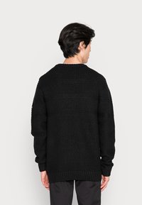 Maglione nero lavorato a maglia con motivo testurizzato, spalle cadenti e orlo a coste. La vista posteriore mostra una vestibilità rilassata e dettagli sottili.