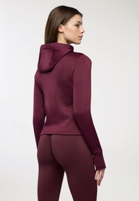 Felpa atletica color rosso amaranto con design aderente, maniche lunghe e cappuccio. Il tessuto appare liscio, abbinandosi a leggings coordinati e aderenti.