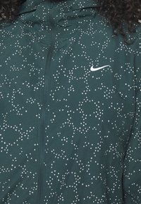 Tyrkysová bunda s předním zipem, která má skvrnitý bílý vzor. Obsahuje kapuci a malé bílé logo Nike na levé straně.