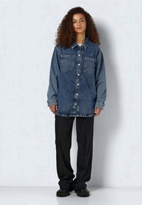 Denim shirt with long sleeves, two chest pockets, and button closure. Distressed blue fabric; paired with loose black trousers and chunky black shoes.