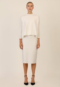 Biały, dziany oversize'owy sweter z prążkowanymi rękawami, zestawiony z dopasowaną białą spódnicą midi. Czarne szpilki w szpic dopełniają stój.