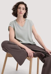 Lichtgroene v-hals t-shirt met korte omgeslagen mouwen, gecombineerd met losse bruine broek, zittend op een houten stoel, met zichtbare bruine franjerap schoenen.