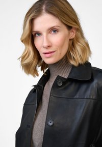 Black leather jacket with a collar and button closure, worn over a light brown ribbed turtleneck. Smooth texture and tailored fit.