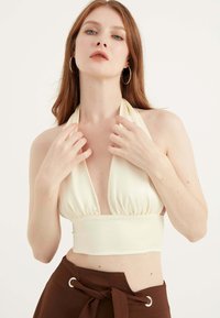 Crème halter neck top met diepe halslijn en geraffineerde bodice, gecombineerd met bruine high-waisted broek met een strik in de taille en oogje-detail.