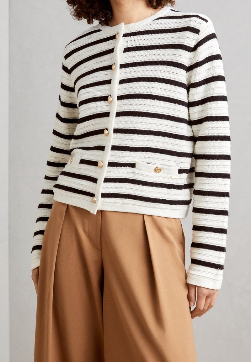 Femme portant un cardigan boutonné rayé blanc et noir avec des boutons dorés et un pantalon large beige sur un fond neutre.