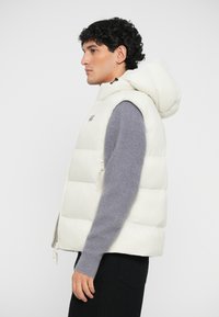 White puffer vest with a hood, featuring quilted sections, a zippered front, and a subtle logo. Worn over a grey sweater.
