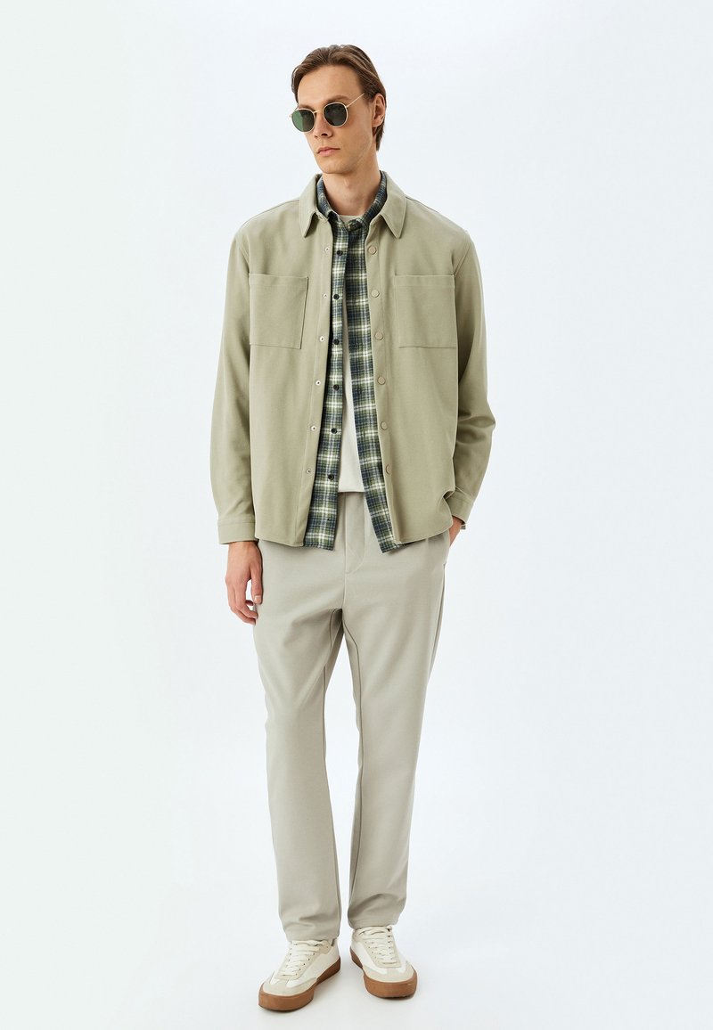 Lysegrøn button-up jakke over en ternet skjorte, parret med beige bukser og hvide sneakers. Model iført grønne solbriller.