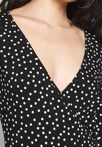 Svart tyg med vita prickar, med en djup V-halsringning och en figurnära design. Slät textur med en lätt stretch.