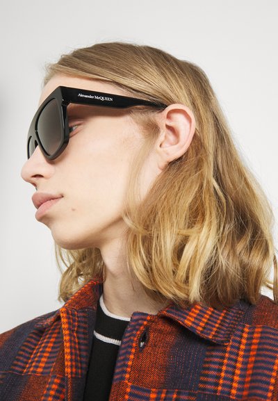Ochelari de soare negri, supradimensionați, cu o formă geometrică și lentile negre. Prezintă logo-ul "Alexander McQueen" pe braț. Purtați cu o jachetă în carouri.