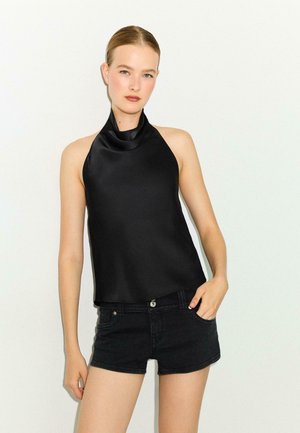 Junge Frau mit zurückgekämmtem Haar, die ein schwarzes Satin-Haltertop und schwarze Jeansshorts vor einem einfarbigen hellen Hintergrund trägt.
