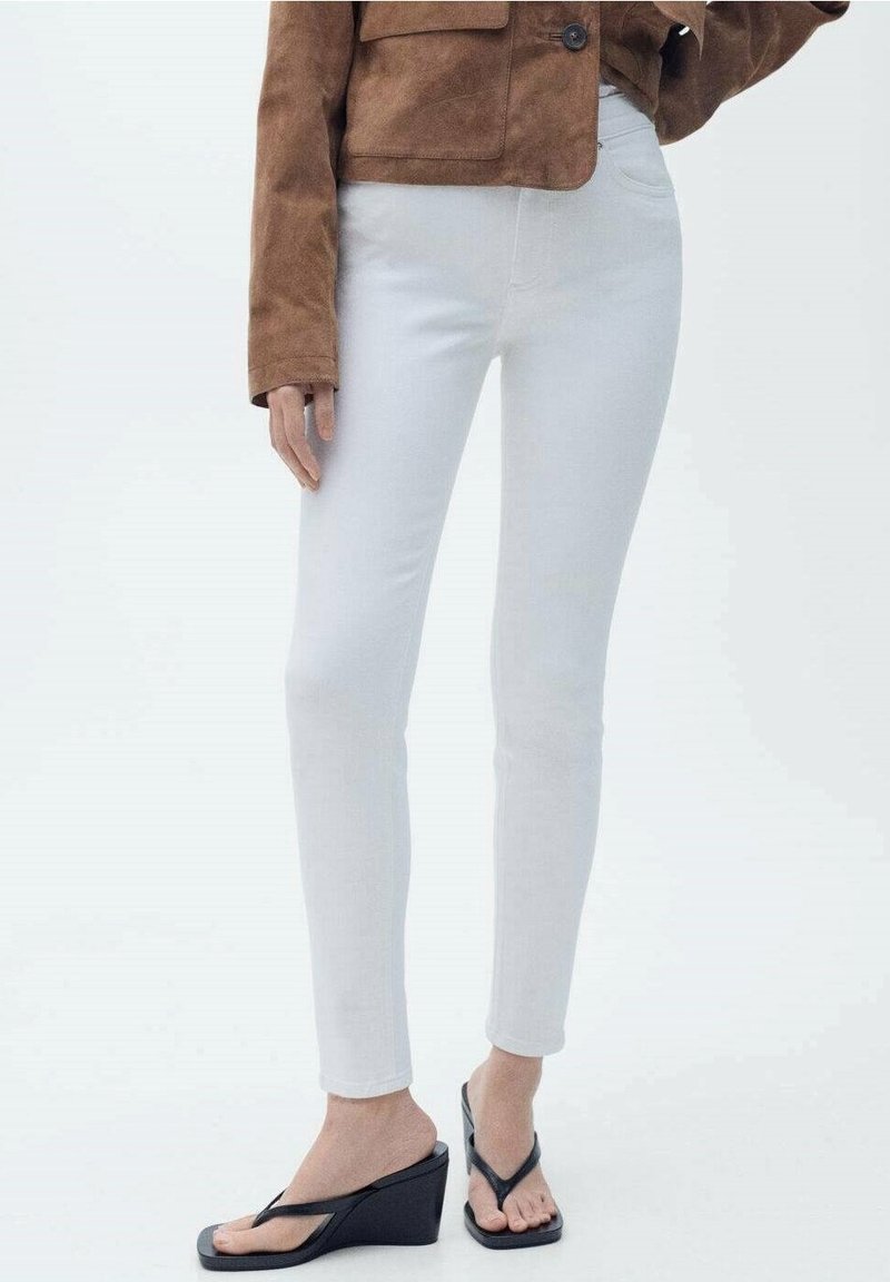 Femme portant un jean skinny blanc, une veste en daim marron et des tongs compensées noires, debout devant un fond clair uni.