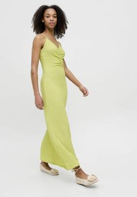 Robe longue ajustée en tissu jaune clair texturé avec un corsage froncé et des fines bretelles, associée à des ballerines beige avec un accent floral.