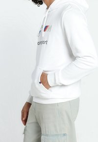 Felpa bianca con cappuccio e tasca frontale, caratterizzata da grafiche del logo BMW M in argento e accenti blu/rossi. Tessuto morbido, polsini e orlo a coste.