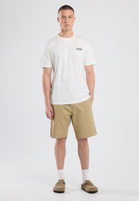 White cotton t-shirt with round neck, tan shorts, white socks, and light brown slip-on shoes, standing on a plain background.