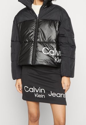 Persoon die een zwarte pufferjas en een zwarte rok draagt, beide met grote witte Calvin Klein Jeans-logo's.