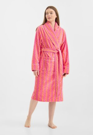 Kvinde står barfodet iført en knælange badekåbe med bølgemønster i pink og orange med bælte og forlommer.