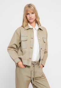 Lys beige denimjakke med to frontlommer, knappelukking og kontraststing. Kombinert med matchende beige bukser.