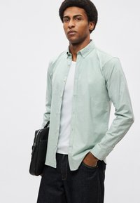 Ανοιχτό πράσινο πουκάμισο με κουμπιά από λείο ύφασμα, με γιακά και εφαρμοστή γραμμή, σε συνδυασμό με σκούρο τζιν παντελόνι και μαύρη τσάντα.