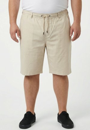 Mann trägt beige Shorts mit Kordelzug, schwarze Sneakers und ein weißes Hemd vor einem einfarbigen weißen Hintergrund.