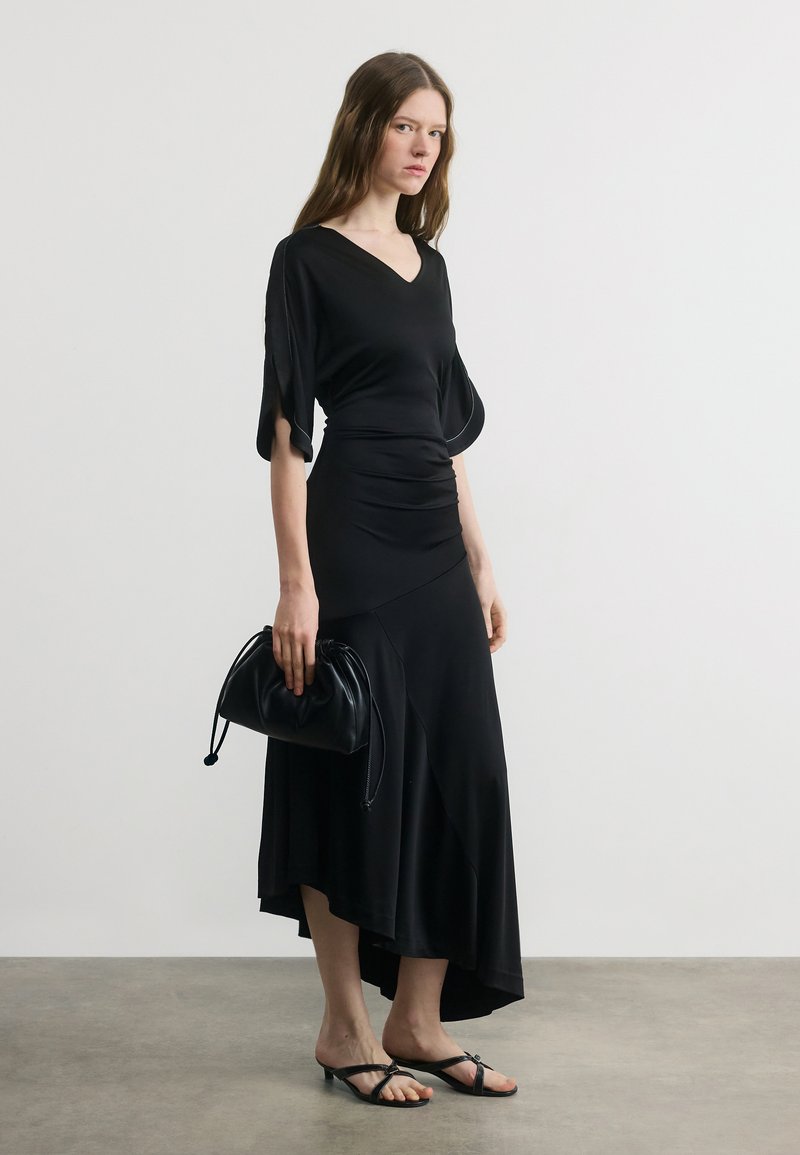 Γυναίκα που φοράει μαύρο midi φόρεμα με μανίκια που κυματίζουν, κρατώντας μαύρη μικρή τσάντα, και φοράει μαύρα πέδιλα με τακούνι, στέκεται σε γκρι πάτωμα.