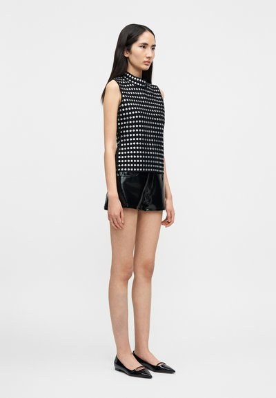 Νεαρή γυναίκα που φορά αμάνικο μαύρο τοπ με λευκούς κύκλους, μαύρα γυαλιστερά σορτς και μαύρα μυτερά παπούτσια, στέκεται μπροστά σε απλό λευκό φόντο.